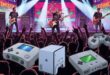 Pra Cego Ver: Uma ilustração que mostra uma banda tocando no palco e abaixo tem o público e a demonstração de alguns consoles de videogame.