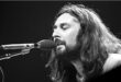 Pra Cego Ver: Foto em preto e branco mostra o músico Rick Davies tocando piano e cantando. Ele está de barba e cabelo comprido