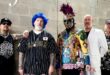 Pra Cego Ver: Foto colorida mostra os integrantes da banda. Eles estão utilizando roupas coloridas e estão em pé do lado um do outro.