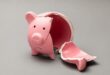 Pra Cego Ver: A imagem é um cofinho de porco rosa quebrado e vazio