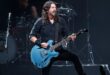 Pra Cego Ver: A foto colorida mostra o músico Dave Grohl em pé, utilizando uma roupa preta e tocando uma guitarra azul clara