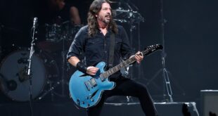 Pra Cego Ver: A foto colorida mostra o músico Dave Grohl em pé, utilizando uma roupa preta e tocando uma guitarra azul clara