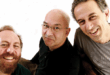 Pra Cego Ver: A foto colorida mostra os integrantes da banda Paralamas do Sucesso rindo e com um fundo branco