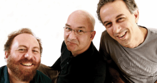 Pra Cego Ver: A foto colorida mostra os integrantes da banda Paralamas do Sucesso rindo e com um fundo branco