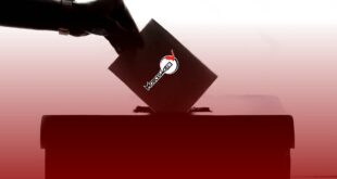 Mão segurando um envelope inclinado para baixo enquanto o insere em uma urna de votação escura. O envelope exibe o logotipo da Morcegão FM. O fundo é totalmente branco com leve degrade vermelho, criando forte contraste com as silhuetas escuras da mão e da urna.